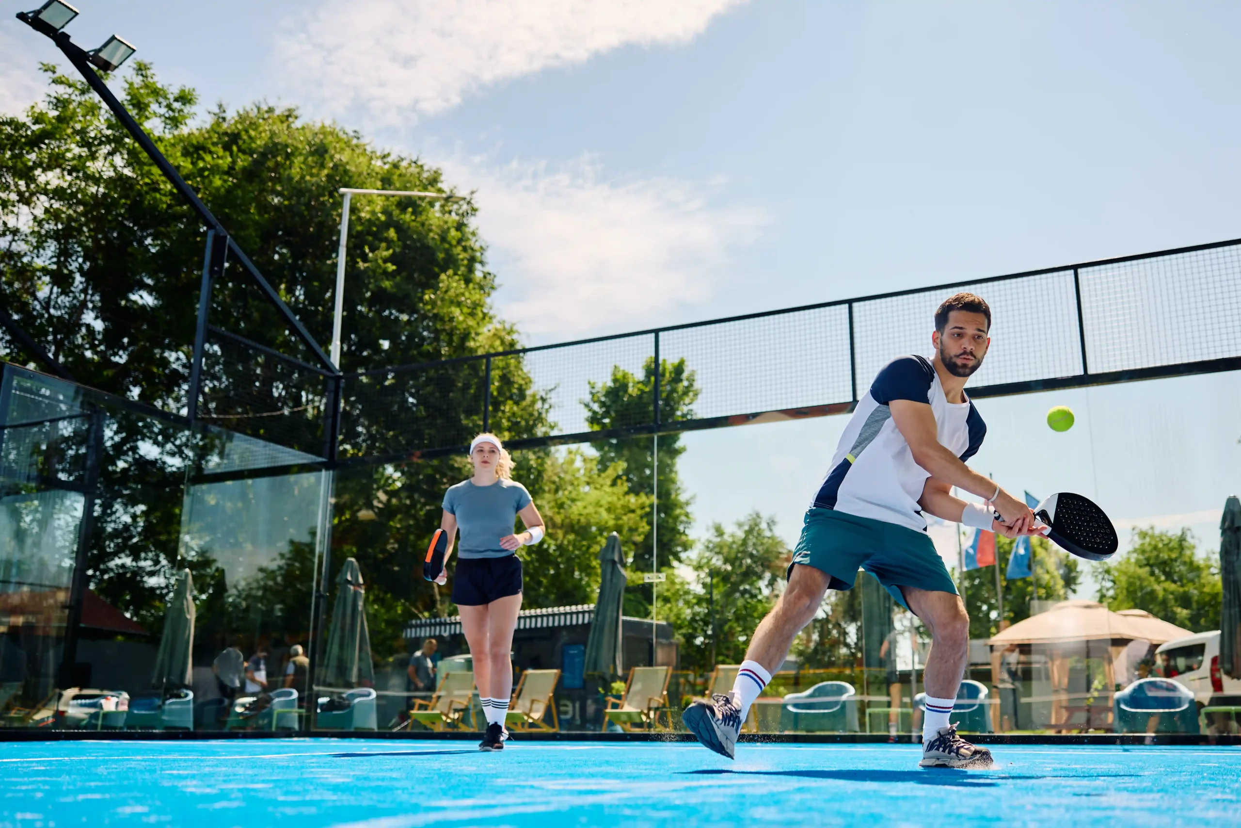 Young man playing padel in Cyprus on a blue outdoor court with partner in the background.
